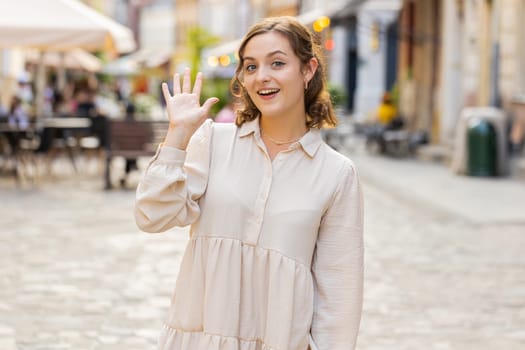 Hello. Young pretty woman smiling friendly at camera, waving hands gesturing hi, greeting or goodbye, welcoming with hospitable expression outdoors. Happy lovely girl walking in urban city town street