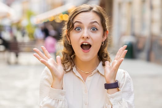 Oh my God, Wow. Young pretty woman looking surprised at camera with big open eyes, shocked by sudden victory, game winning, lottery goal achievement, good news outdoors. Lovely girl in city street