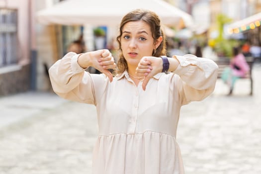 Dislike. Upset young pretty woman showing thumbs down sign gesture, expressing discontent, disapproval, dissatisfied bad work, mistake. Displeased lovely girl walking in urban city street outdoors