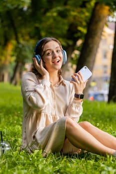 Happy relaxed young woman in wireless headphones choosing, listening favorite energetic disco rock n roll music in smartphone dancing outdoor. Girl sitting on grass in urban sunset city park. Vertical