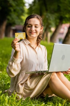 Young woman using credit bank card and laptop computer while transferring money, purchases online shopping, order food delivery. Girl sitting on grass in urban sunset city park background. Vertical