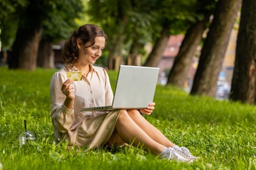 Portrait of young woman using credit bank card and laptop computer while transferring money, purchases online shopping, order food delivery. Girl sitting on grass in urban sunset city park background