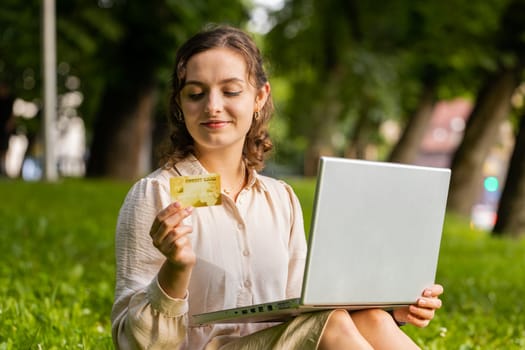 Portrait of young woman using credit bank card and laptop computer while transferring money, purchases online shopping, order food delivery. Girl sitting on grass in urban sunset city park background