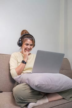 Young beautiful asian woman relaxing and listening to music using headphones, she is lying in sofa. Young woman enjoying at home using laptop...