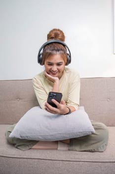 A charming young Asian woman in casual clothes enjoys the music on her headphones while sitting on a sofa in her living room...