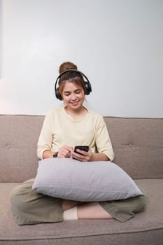 A charming young Asian woman in casual clothes enjoys the music on her headphones while sitting on a sofa in her living room...