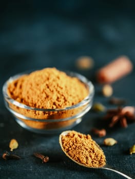 Indian or pakistani masala powder in spoon and glass jar. Homemade dry curry garam masala mix spices blend on dark gray and blue background. Vertical
