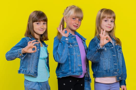 Okay. Teenage girls. Little children sisters looking approvingly at camera showing ok gesture, like sign positive, approve something good. Three siblings kids isolated on studio yellow background