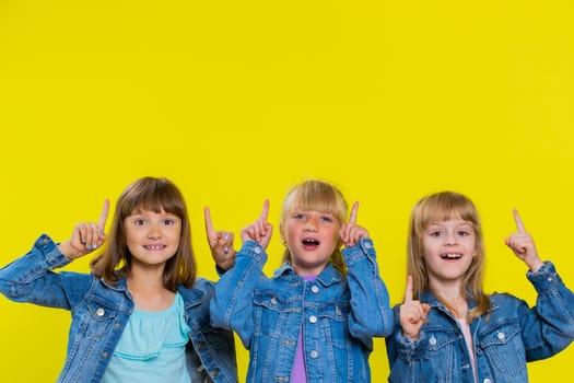 Teenage girls. Little children sisters showing thumbs up and pointing empty place, advertising area for commercial text, copy space for goods promotion. Three siblings kids on studio yellow background