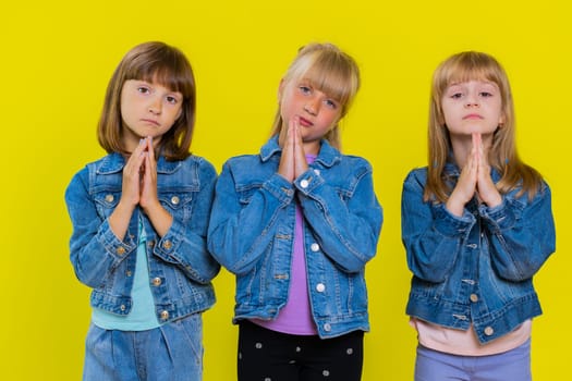 Please, God help. Teenage girls praying, making wish, asking with hopeful imploring expression, begging apology, forgiveness. Little children sisters. Three siblings kids isolated on yellow background