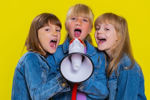 Teenage girls talking with megaphone, proclaiming news, loudly announcing advertisement, warning using loudspeaker to shout speech. Little children sisters. Three siblings kids on yellow background