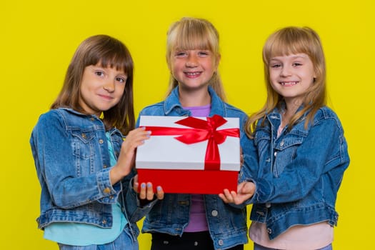 Teenage girls presenting birthday gift box with red ribbon stretches out hands offer wrapped present celebrating party, advertising holidays sale discount. Little children sisters. Three siblings kids