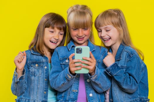 Teenage girls use mobile smartphone typing browsing say Wow yes found out great big win good news celebrate, playing online game. Little children sisters. Three siblings kids on yellow background