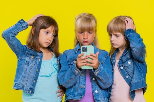 Teenage girls use smartphone typing browsing, loses becoming surprised sudden lottery results, bad news, fortune loss, fail. Little children sisters. Three siblings kids isolated on yellow background