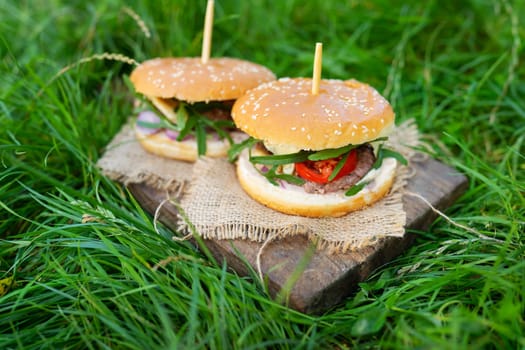 Delicious fresh burgers served on wooden boards, photo outdoors on green grass. Close-up