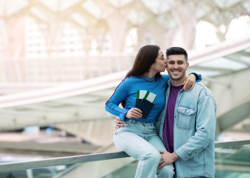 Great Travel Offer. Joyful Woman Kissing Her Husband Holding Passports With Boarding Pass Tickets Posing In Modern Airport Indoors. Couple Celebrating Vacation. Empty Space For Text