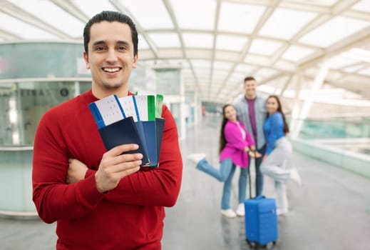 Great Travel Offer. Tourist Man Showing Passports And Boarding Passes Posing With Group Of Friends At Airport Indoor. Tour Agent Advertising Transportation Service. Selective Focus