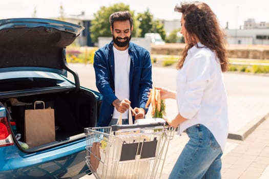 Young middle eastern spouses packing groceries into car trunk outdoors, standing on parking near shopping mall. Sales in supermarket