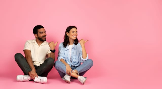 Summer deal. Curious happy beautiful young couple man and woman wearing casual summer outfits sitting on floor over pink studio background, pointing with thumbs at copy space, panorama