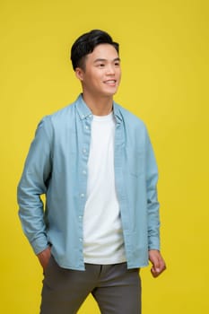  smiling young handsome Asian man standing with one hand in pocket isolated on yellow studio background