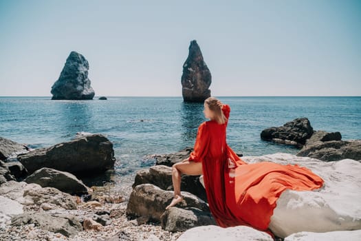 Woman travel sea. Young Happy woman in a long red dress posing on a beach near the sea on background of volcanic rocks, like in Iceland, sharing travel adventure journey