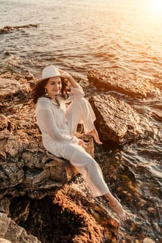 woman sea sunset. woman in a white pantsuit and hat is sitiing on the beach enjoying the sea. Happy summer holiday.