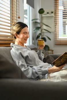 Relaxed young woman drinking hot coffee and reading book, spending free time in Sunday morning at home.