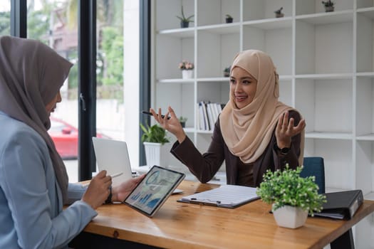 Young muslim female business worker discussing with friends sitting together, working with tablet and financial documents at desk.
