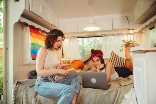 Portrait of a cute lesbian couple. Two girls spend time tenderly together watching movie on laptop in a camper trailer with LGBT flag on the wall. Love and attitude. LGBT concept. High quality photo