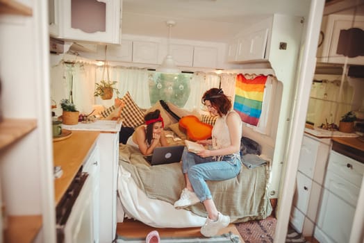 Portrait of a cute lesbian couple. Two girls spend time tenderly together watching movie on laptop in a camper trailer with LGBT flag on the wall. Love and attitude. LGBT concept. High quality photo