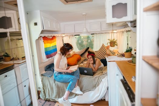 Portrait of a cute lesbian couple. Two girls spend time tenderly together watching movie on laptop in a camper trailer with LGBT flag on the wall. Love and attitude. LGBT concept. High quality photo