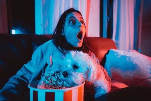 Close up portrait of Beautiful young girl at home in a dark room with a popcorn bucket playing with her dog and sits in front of a monitor or TV watching movie. Crazy emotions. High quality photo
