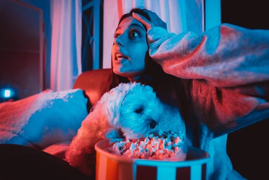 Beautiful young girl at home in a dark room with a popcorn bucket playing with her dog and sits in front of a monitor or TV watching movie. Crazy emotions. High quality photo