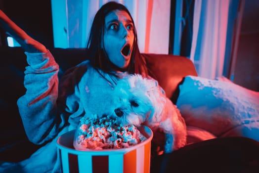 Close up portrait of Beautiful young girl at home in a dark room with a popcorn bucket playing with her dog and sits in front of a monitor or TV watching movie. Crazy emotions. High quality photo