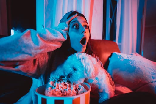 Close up portrait of Beautiful young girl at home in a dark room with a popcorn bucket playing with her dog and sits in front of a monitor or TV watching movie. Crazy emotions. High quality photo