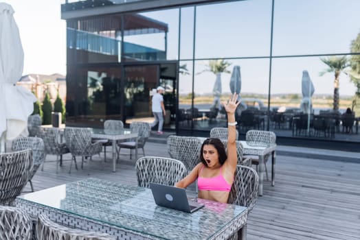 Cute young woman with crazy emotions sitting on terrace, while using laptop for work at summer day
