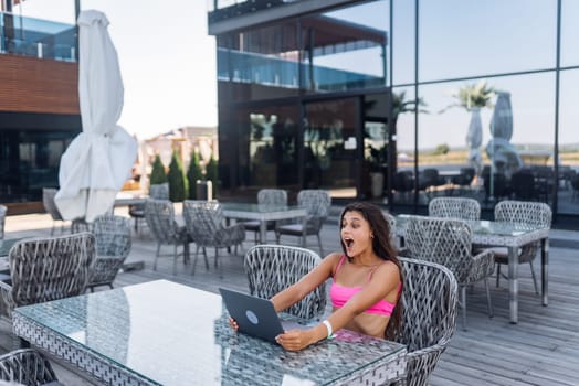 Cute young woman with crazy emotions sitting on terrace, while using laptop for work at summer day
