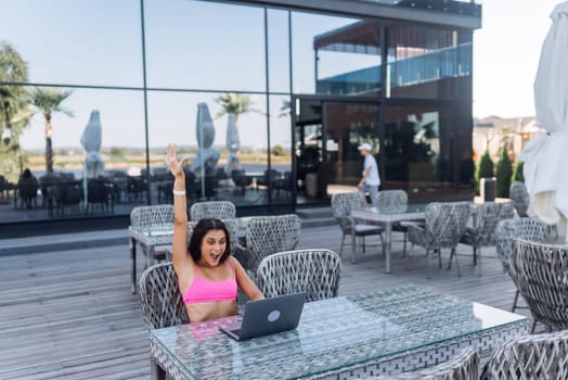 Cute young woman with crazy emotions sitting on terrace, while using laptop for work at summer day