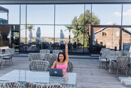 Cute young woman with crazy emotions sitting on terrace, while using laptop for work at summer day