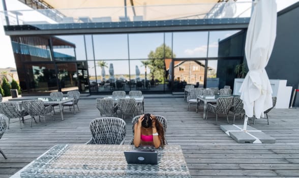 Cute young woman sitting on terrace, while using laptop for work at summer day