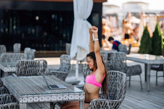 Cute young woman sitting on terrace, happy with hands up winner at summer day