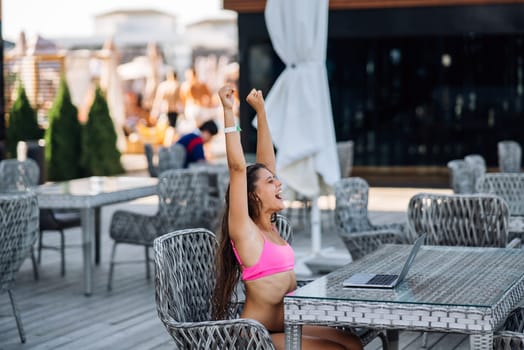 Cute young woman sitting on terrace, happy with hands up winner at summer day