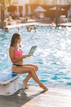 Cute young woman sitting in front of the pool, working with a laptop at summer day
