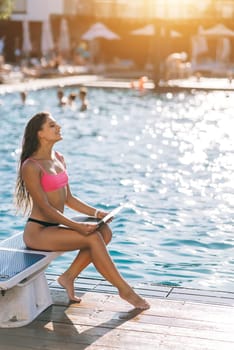 Cute young woman sunbathing sitting in front of the pool, working with a laptop at summer day