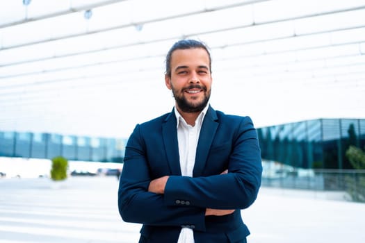 Confidence successful businessman or worker in suit with beard standing business district glass building arm crossed looking at camera and smile. Hispanic male business person portrait. Copy space