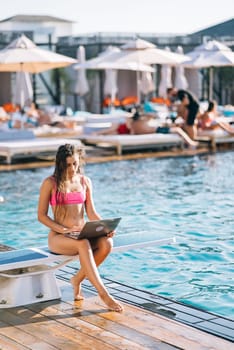 Cute young woman sunbathing sitting in front of the pool, working with a laptop at summer day