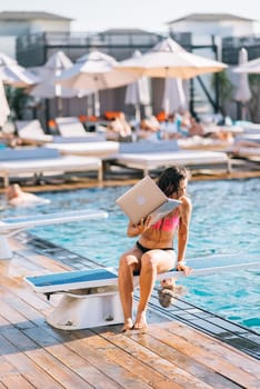 Cute young woman sunbathing sitting in front of the pool, working with a laptop at summer day