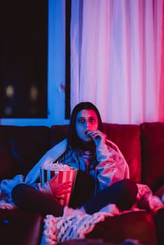 Beautiful young girl eating popcorn watching movie at home in a dark room with a popcorn bucket and playing with her dog and sits in front of a monitor or TV watching movie. Crazy emotions. High quality photo