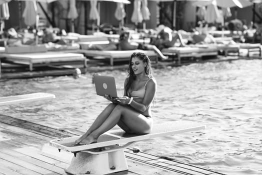 Cute young woman sunbathing sitting in front of the pool, working with a laptop at summer day. Black and white photo