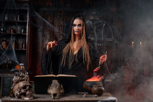 Halloween, witch use magic book and cauldron prepare poison or love potion. Black magic female wizard in dark room cage spider web human skull. Necromancer cooking magic potion. voodoo magic